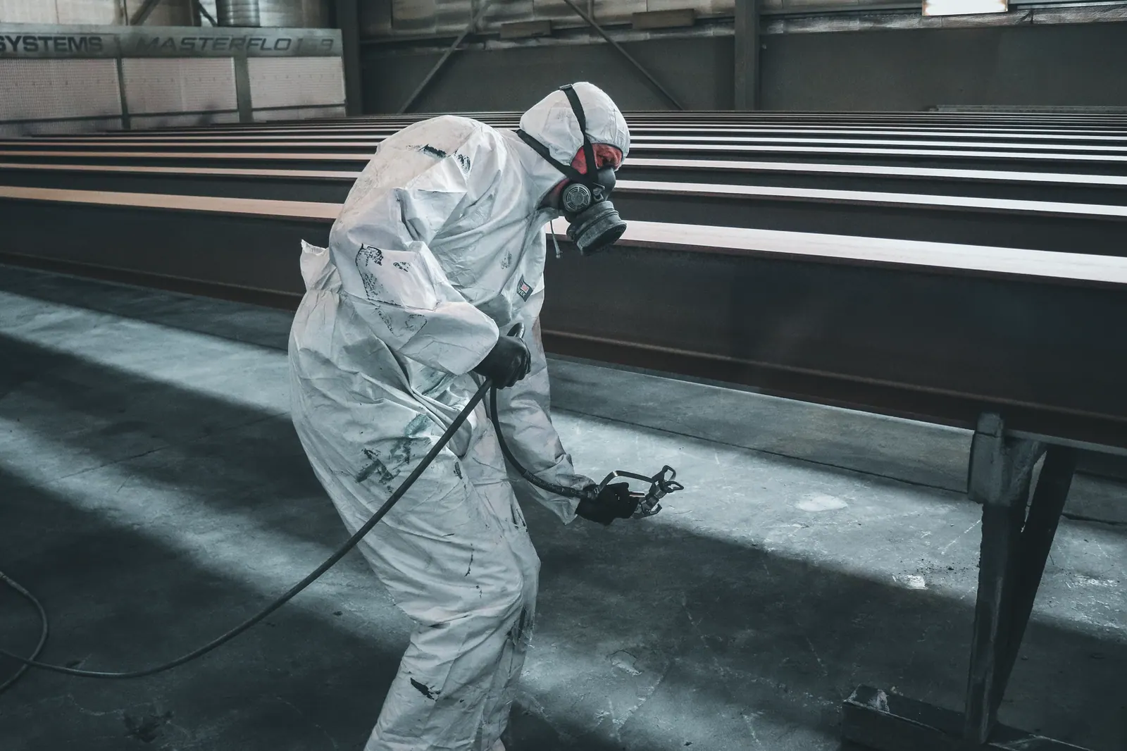 Worker in protective gear spray painting metal beams inside a factory setting.